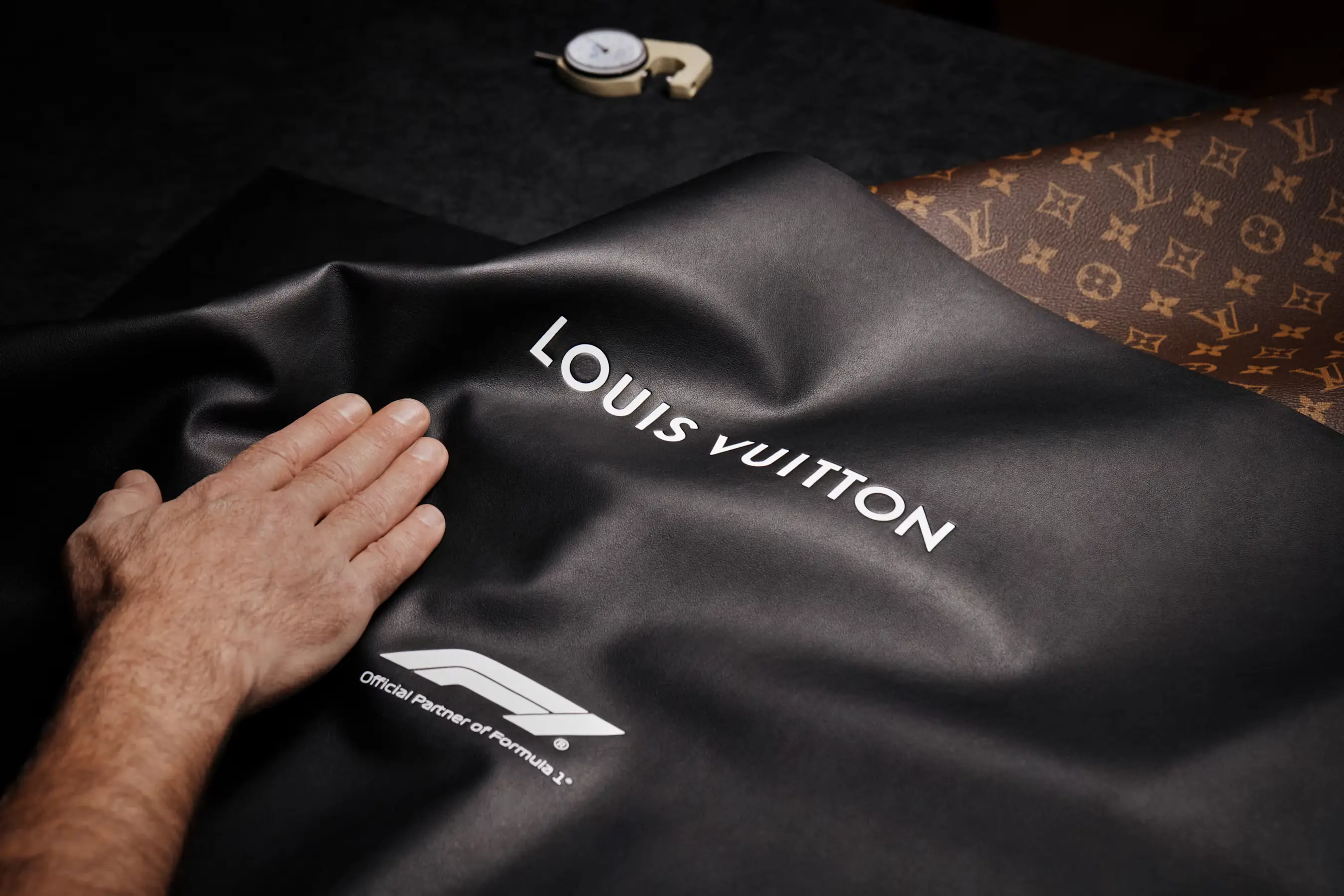 Black Louis Vuitton leather embossed with the Louis Vuitton logo and "Official Partner of Formula 1" emblem, shown beside the brand's monogram canvas.