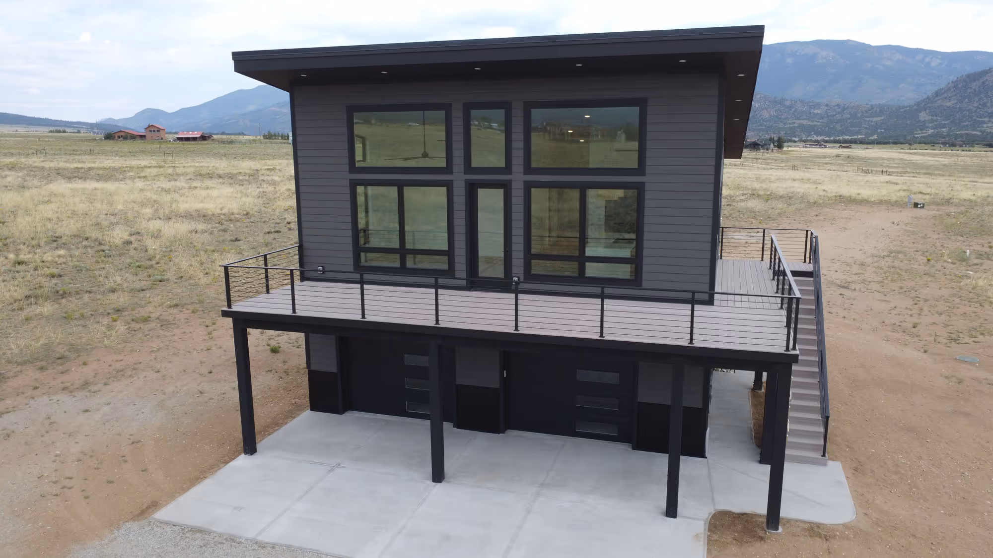 A modern two-story home with dark siding and expansive windows, situated in a wide open field with mountains in the background. The home features a wraparound deck with cable railing and a concrete driveway beneath.