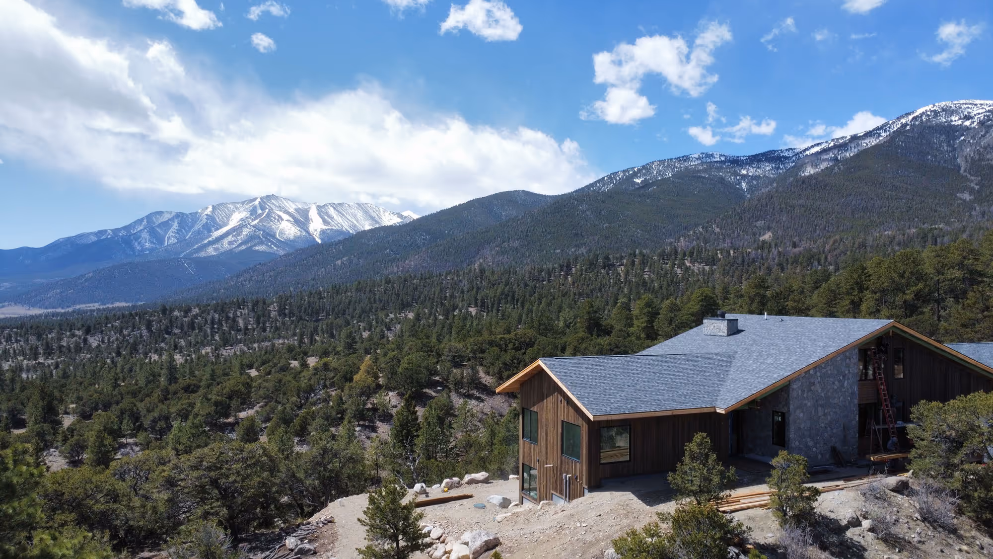 A cabin-style home under construction nestled in a mountainous forest landscape, with snow-capped peaks in the distance and clear blue skies above.
