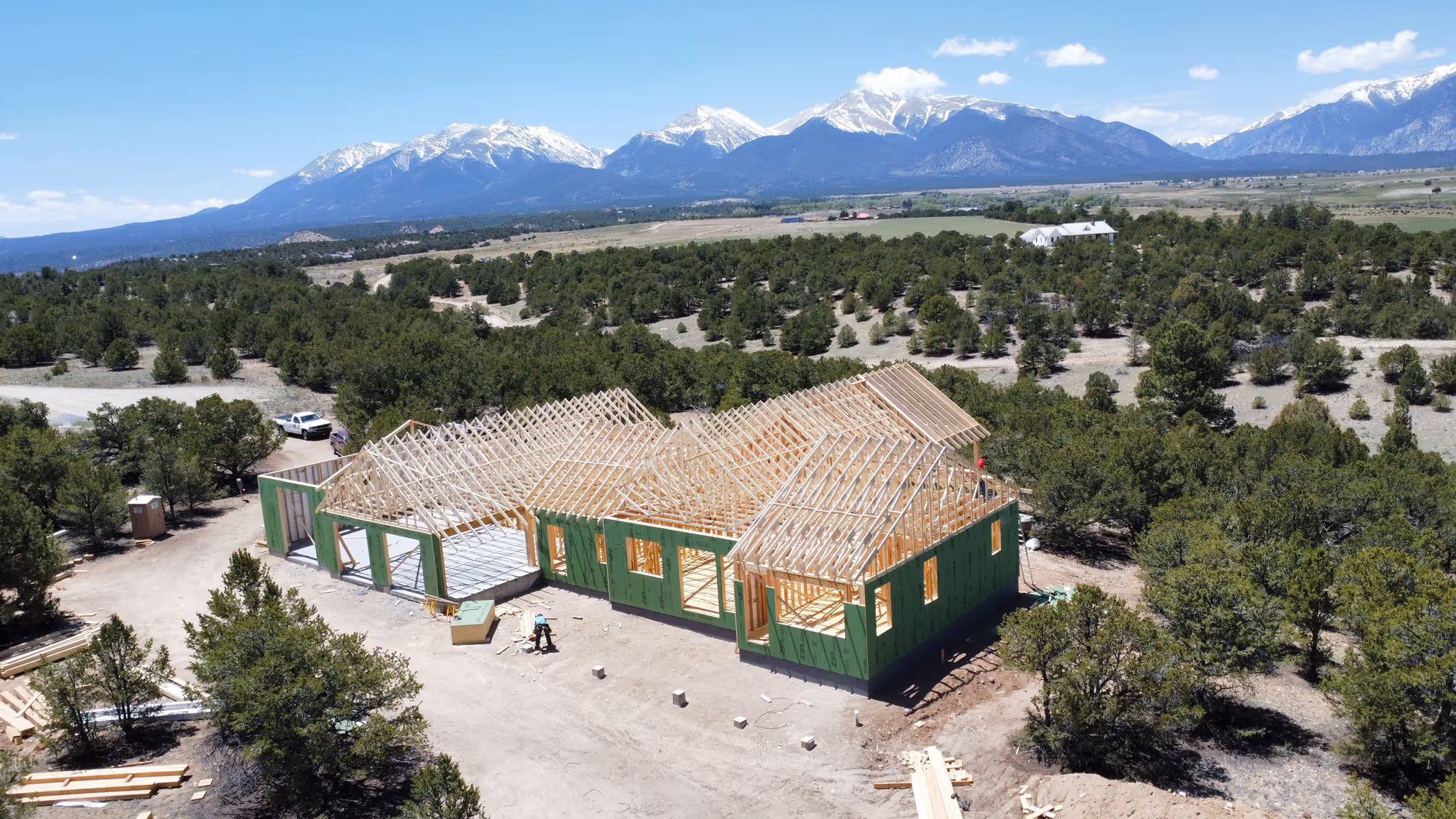 A home in the framing stage with green sheathing and roof trusses in place, set in a rural landscape with distant snow-covered mountains.