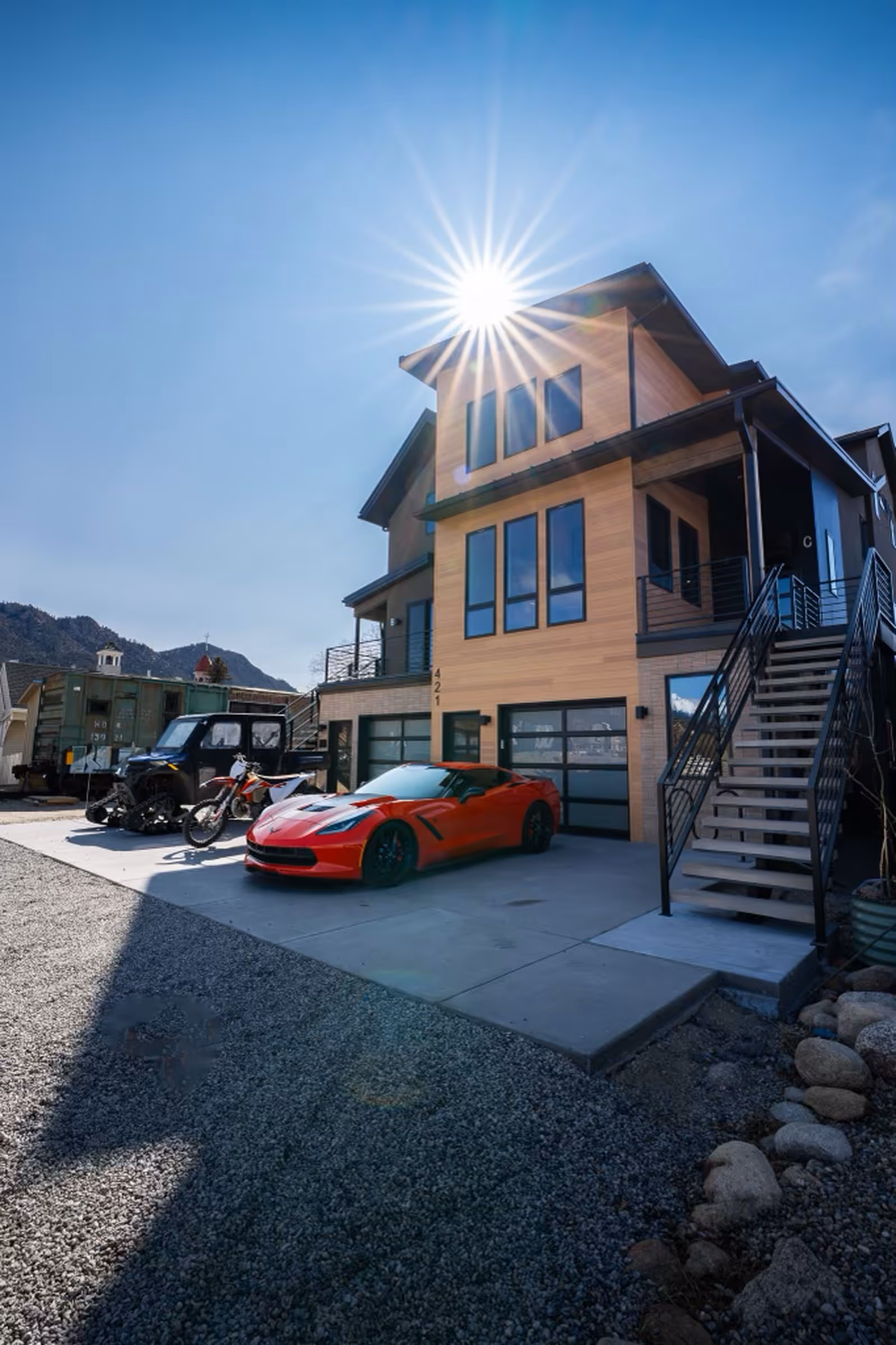 A striking modern mountain home with wood and black siding, flanked by a red Corvette, dirt bikes, and a utility vehicle, with sun rays beaming over the roof.