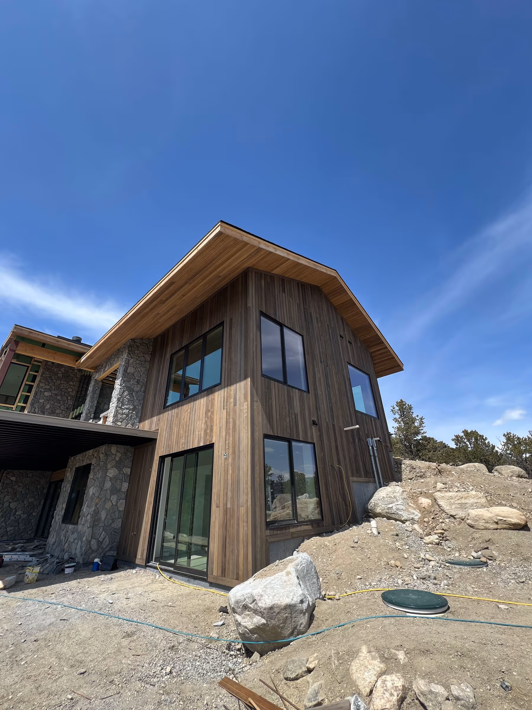 Close-up of a corner section of a home with vertical wood siding, large glass windows, stone walls, and natural boulders in the landscaping.