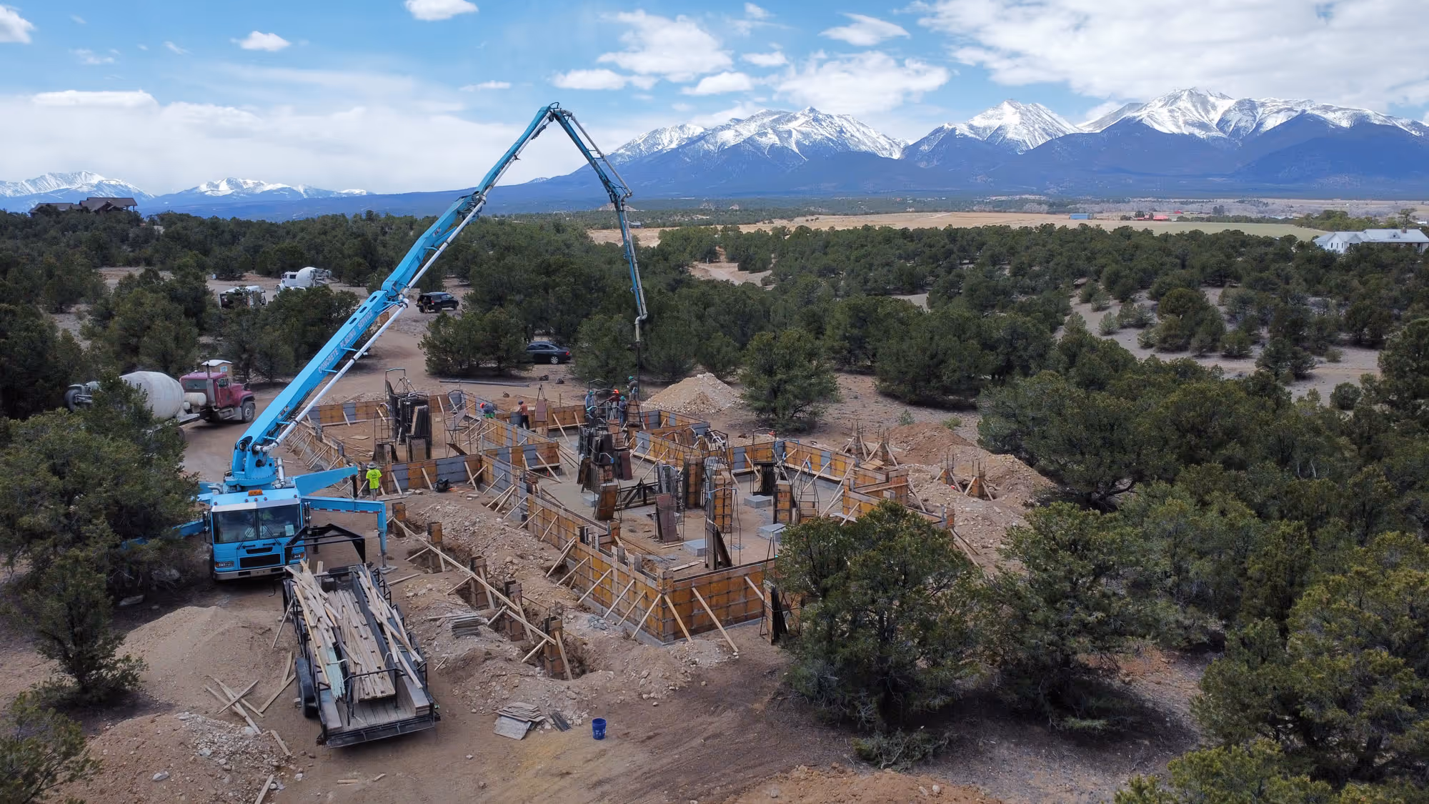 A concrete foundation pour in progress at a wooded mountain construction site, with workers, a concrete pump truck, and framing forms visible.