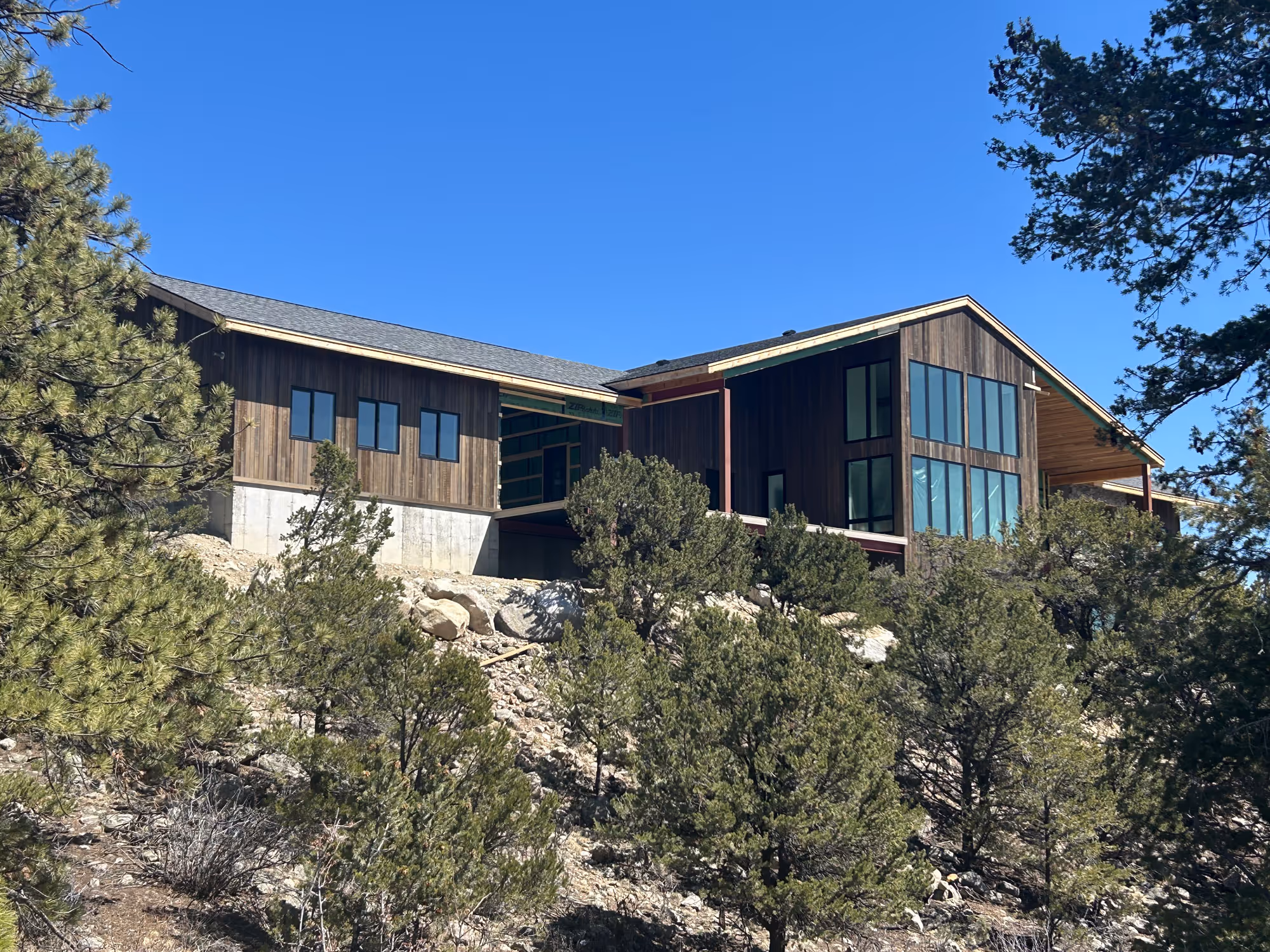 A nearly completed mountain home with wood siding and large windows, perched on a slope with trees and clear blue skies in the background.