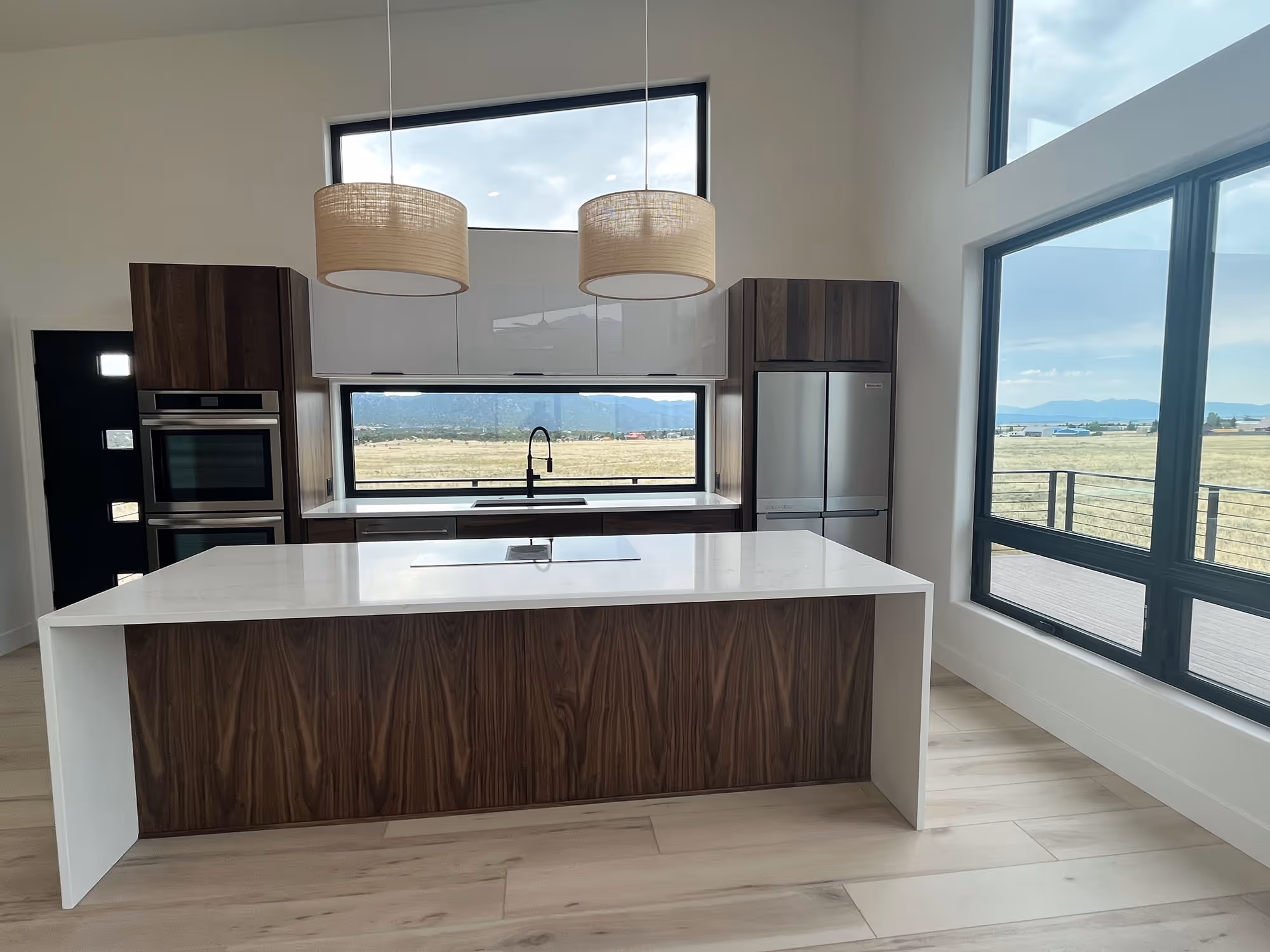 A modern kitchen interior with tall windows, a large island with a waterfall countertop, walnut cabinetry, and mountain views beyond the glass.