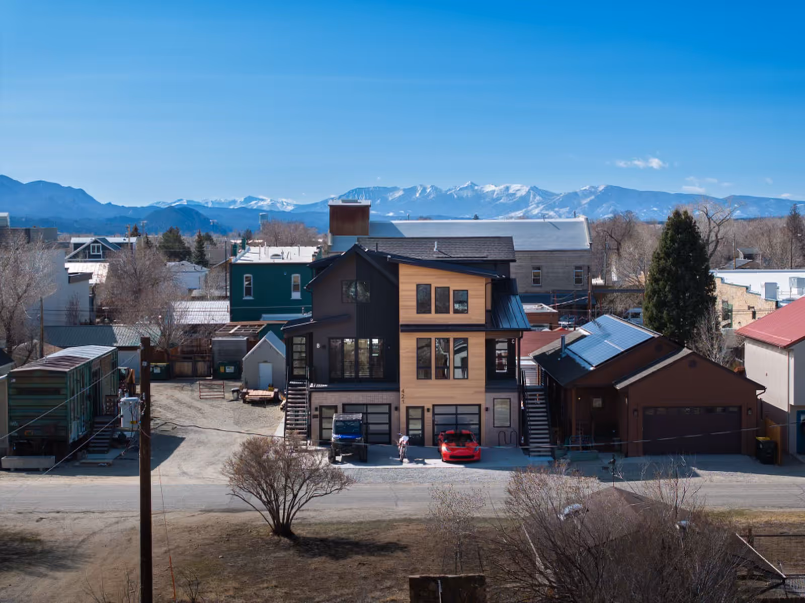 Modern three-story home with wood and dark siding, large windows, and a red sports car in the driveway, set in a small town with mountain views and clear blue skies in the background.