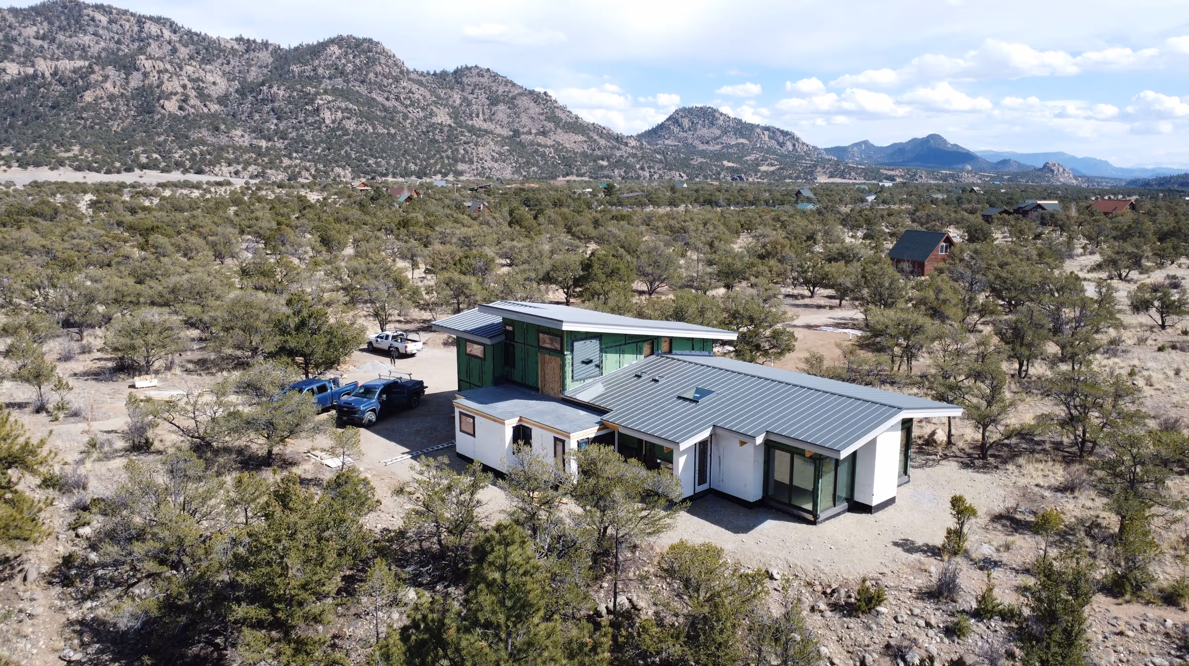 Modern home under construction with green sheathing and a gray metal roof, surrounded by trees and mountains, with multiple trucks parked nearby.