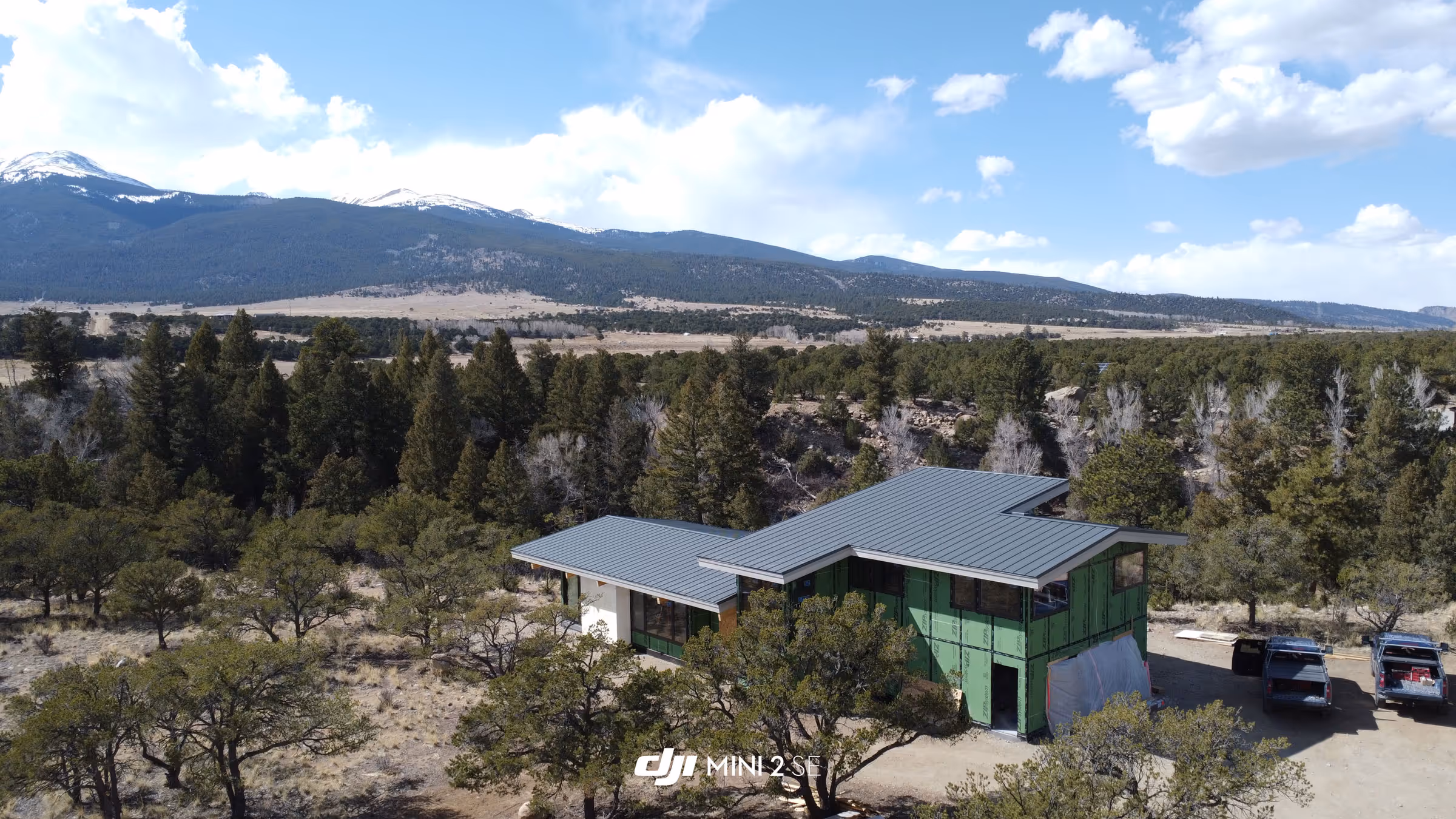 Two-story house under construction with a gray metal roof and green exterior sheathing, set in a wooded landscape with mountain views in the background.
