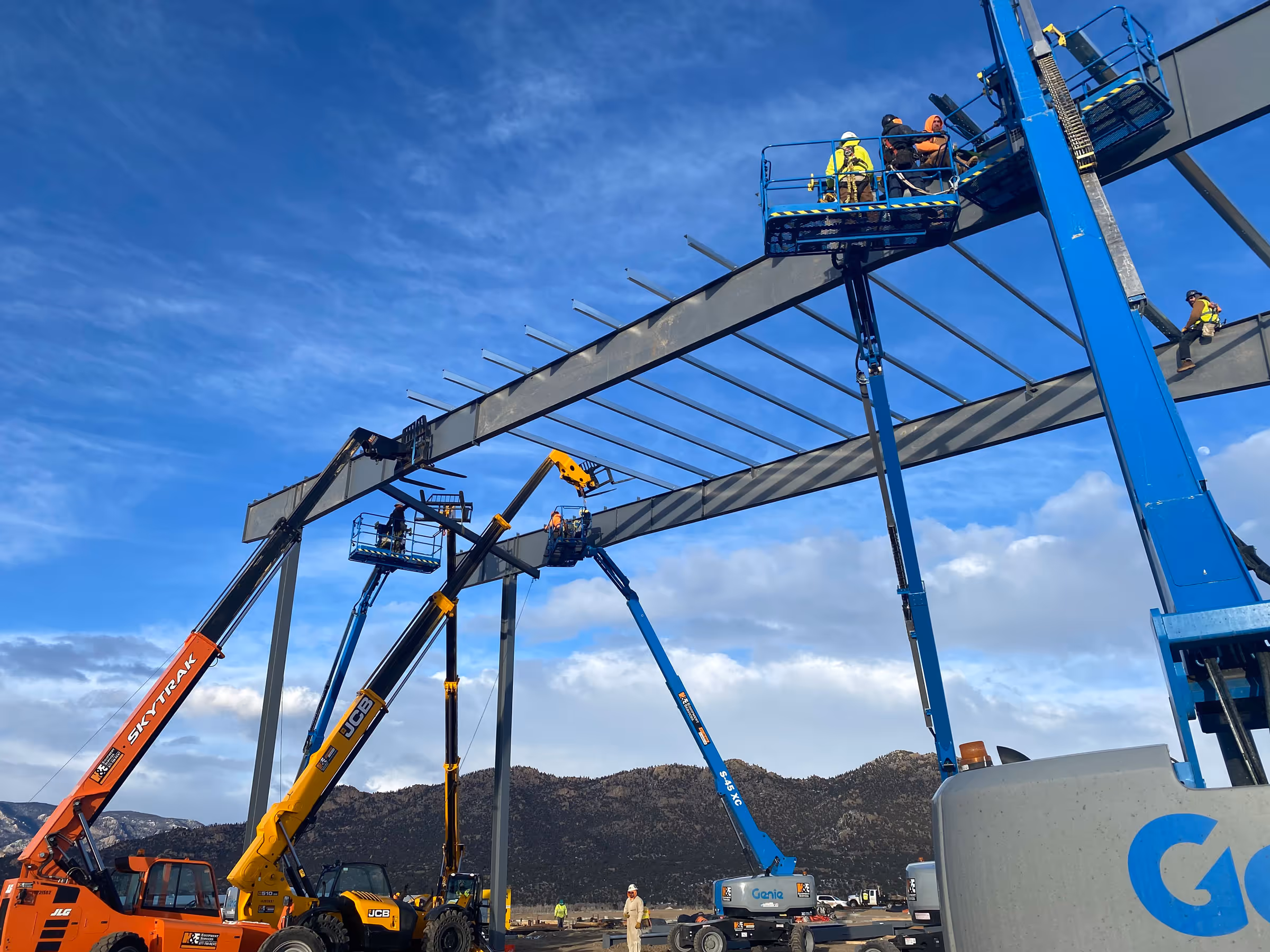 Construction workers using multiple boom lifts to install large steel beams on a commercial building frame against a blue sky and mountain backdrop.