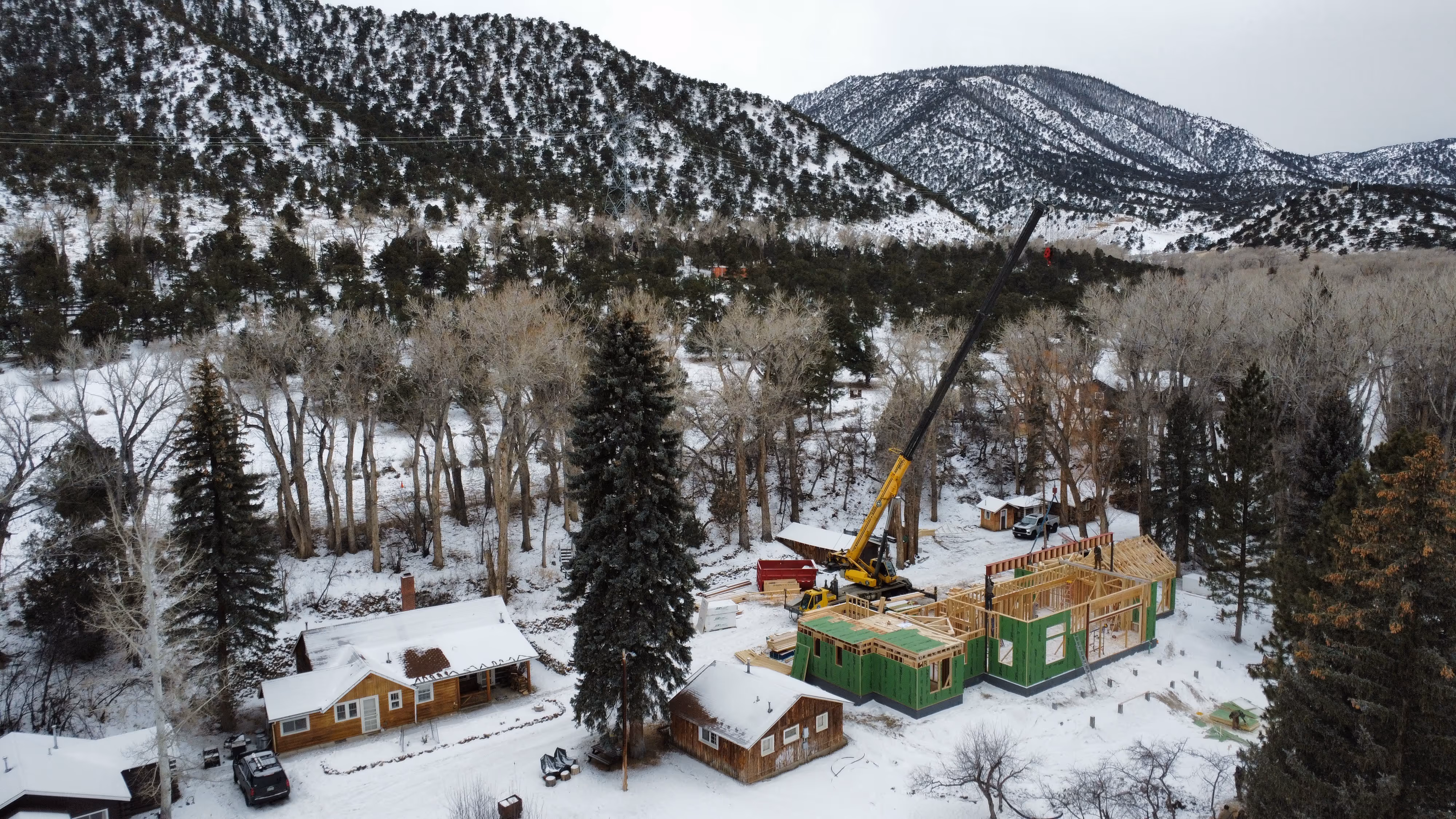 Snow-covered home construction site in a forested valley with a crane lifting roof components and framing underway, surrounded by rustic cabins and tall pines.