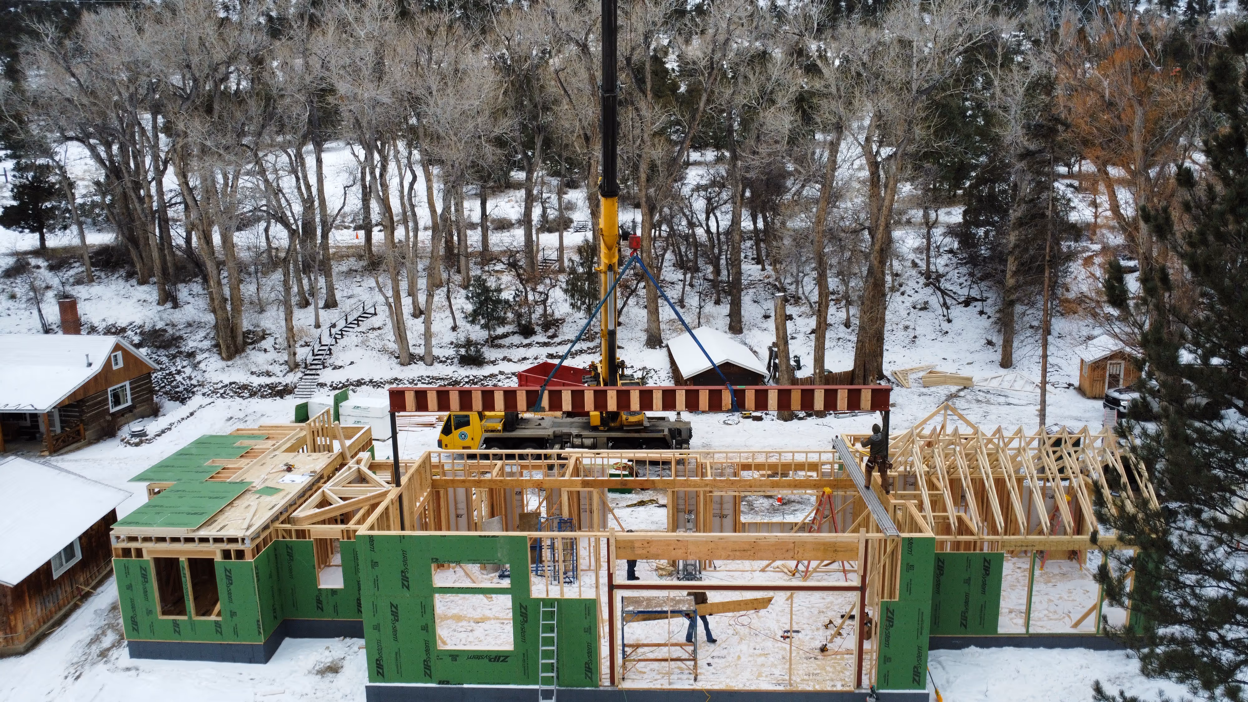 A crane lowers a large steel beam onto a partially framed green-sheathed house during winter, surrounded by snow, trees, and nearby cabins.