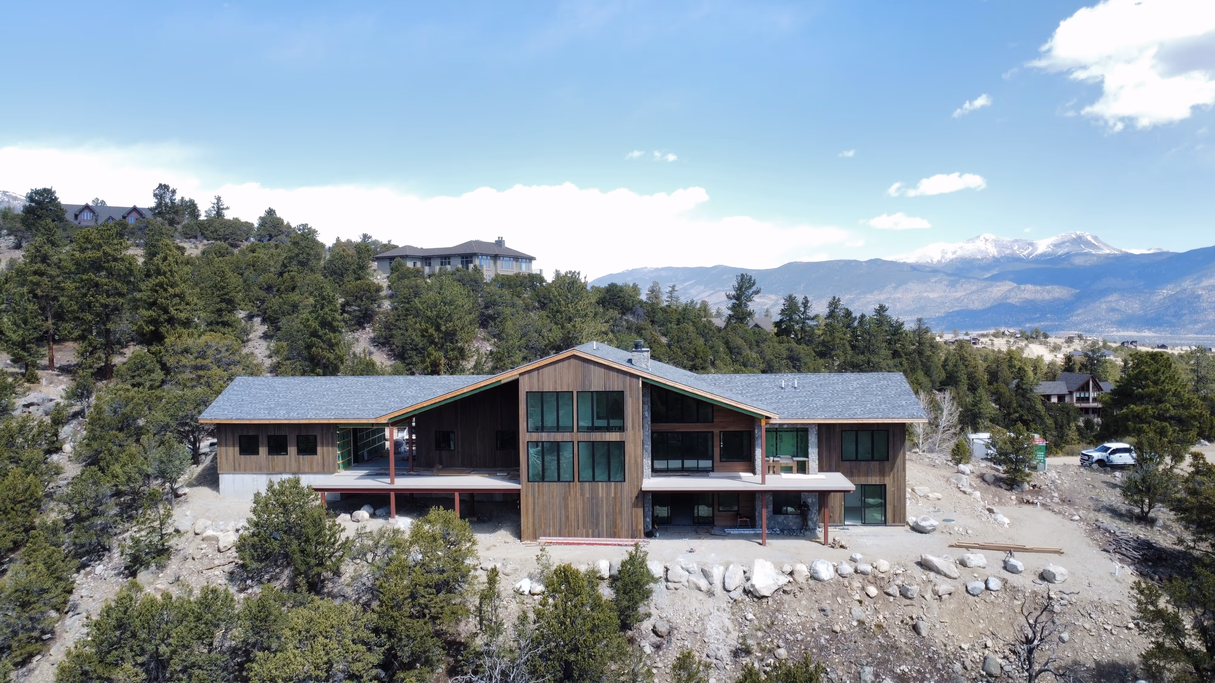 Large modern mountain home with wood and stone exterior, perched on a slope with expansive windows and distant views of snowy peaks.