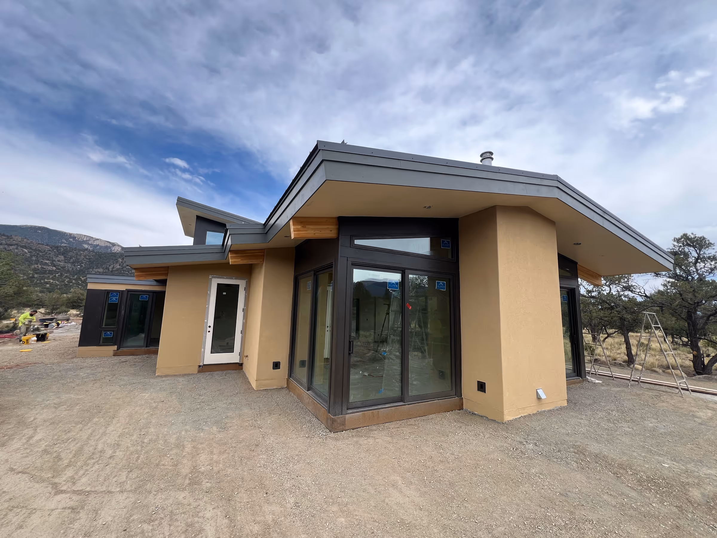 Nearly completed modern home with tan stucco siding, sharp rooflines, and floor-to-ceiling windows, set on a dirt lot with mountainous landscape in the distance.