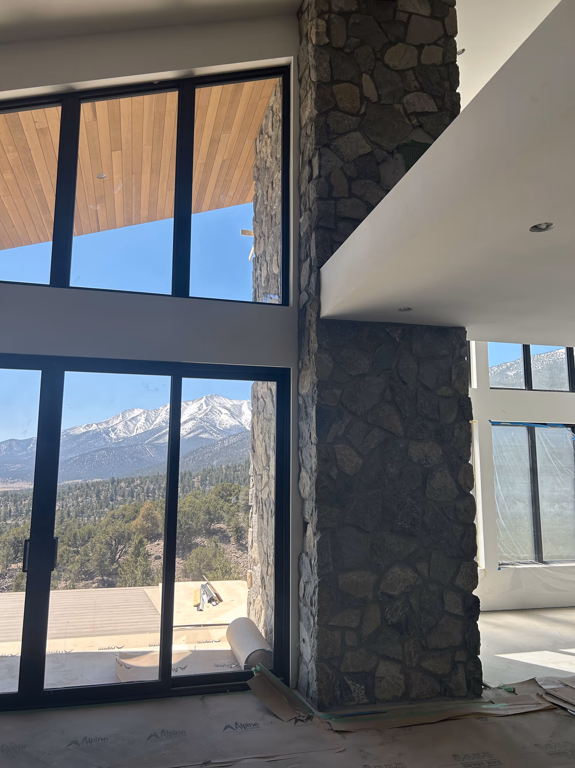 View from inside a mountain home with tall stone columns and large windows framing a snow-capped mountain range beyond the deck.