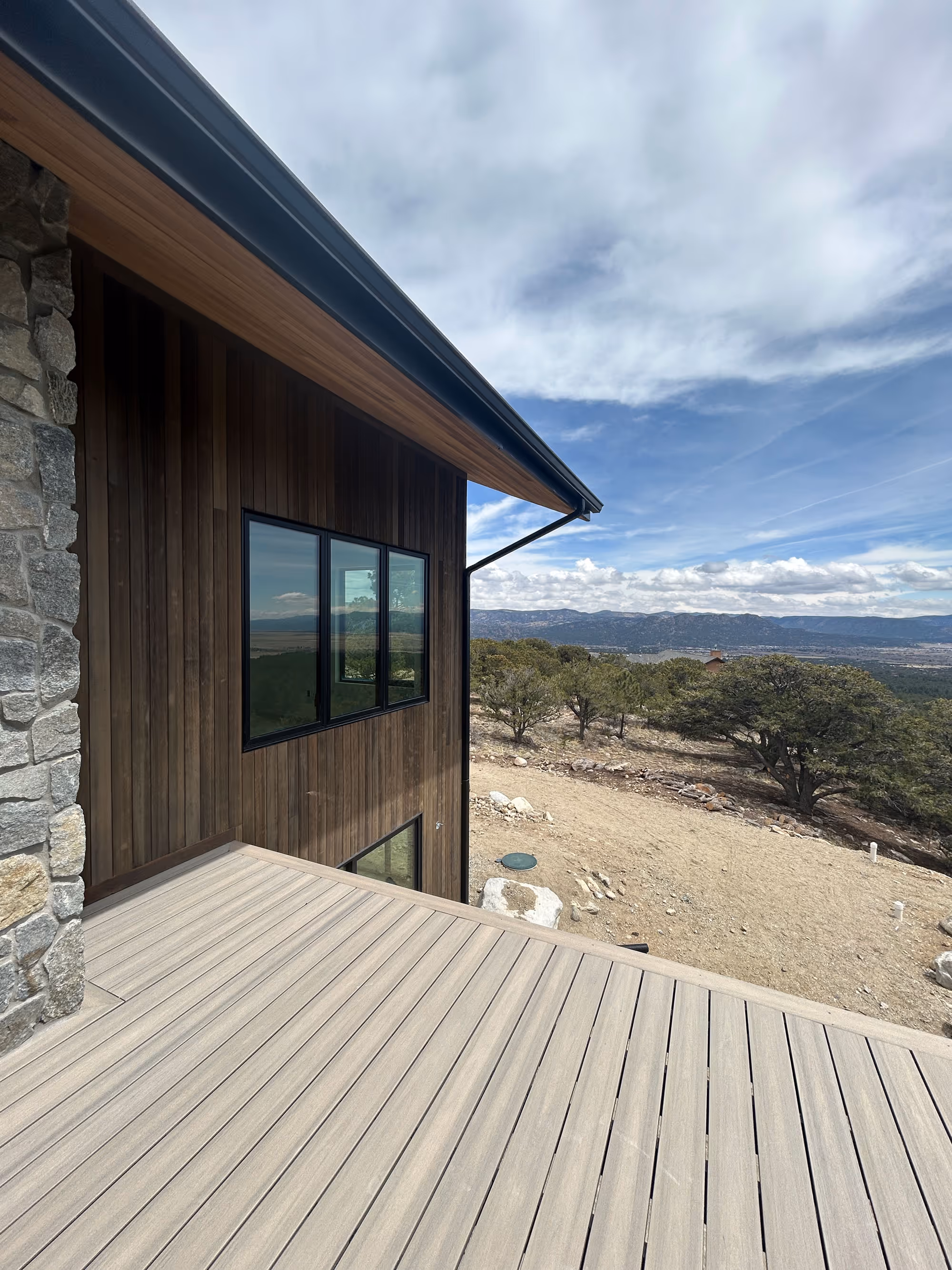 Exterior corner of a mountain home with vertical wood siding and stone accents, looking out from a composite deck toward rolling hills and cloudy sky.