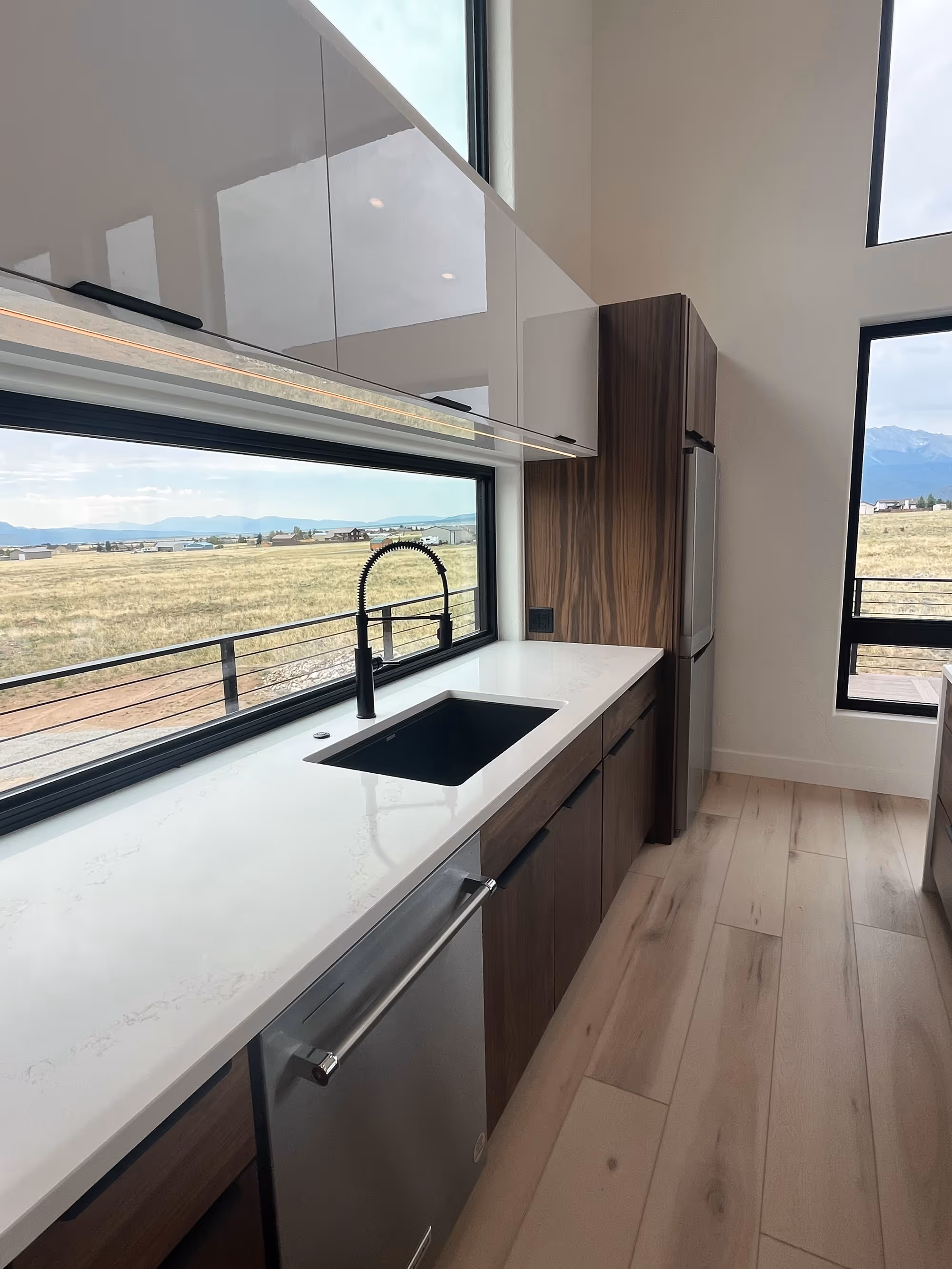 Modern kitchen with clean white countertops, dark wood lower cabinets, black sink and faucet, and expansive windows offering a scenic mountain view.