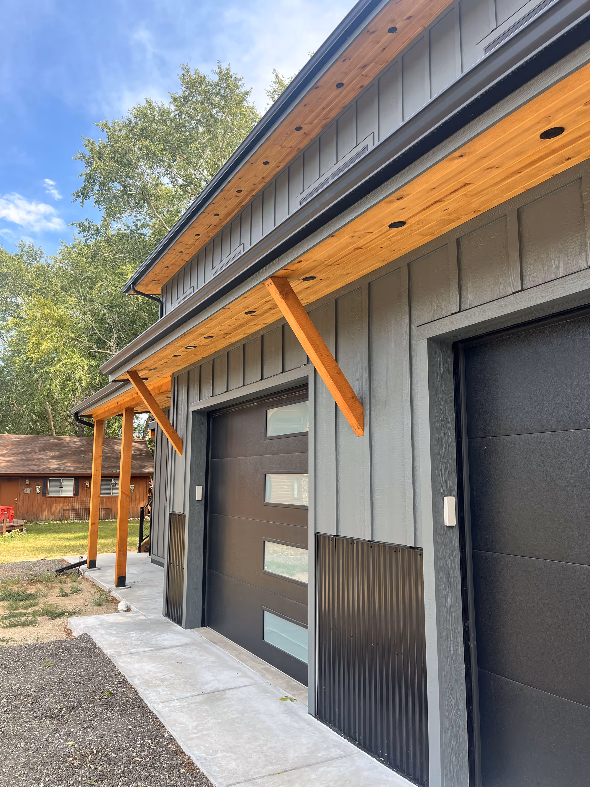 Contemporary garage exterior featuring black doors with glass inserts, vertical gray siding, and natural wood accents beneath the roof overhang.