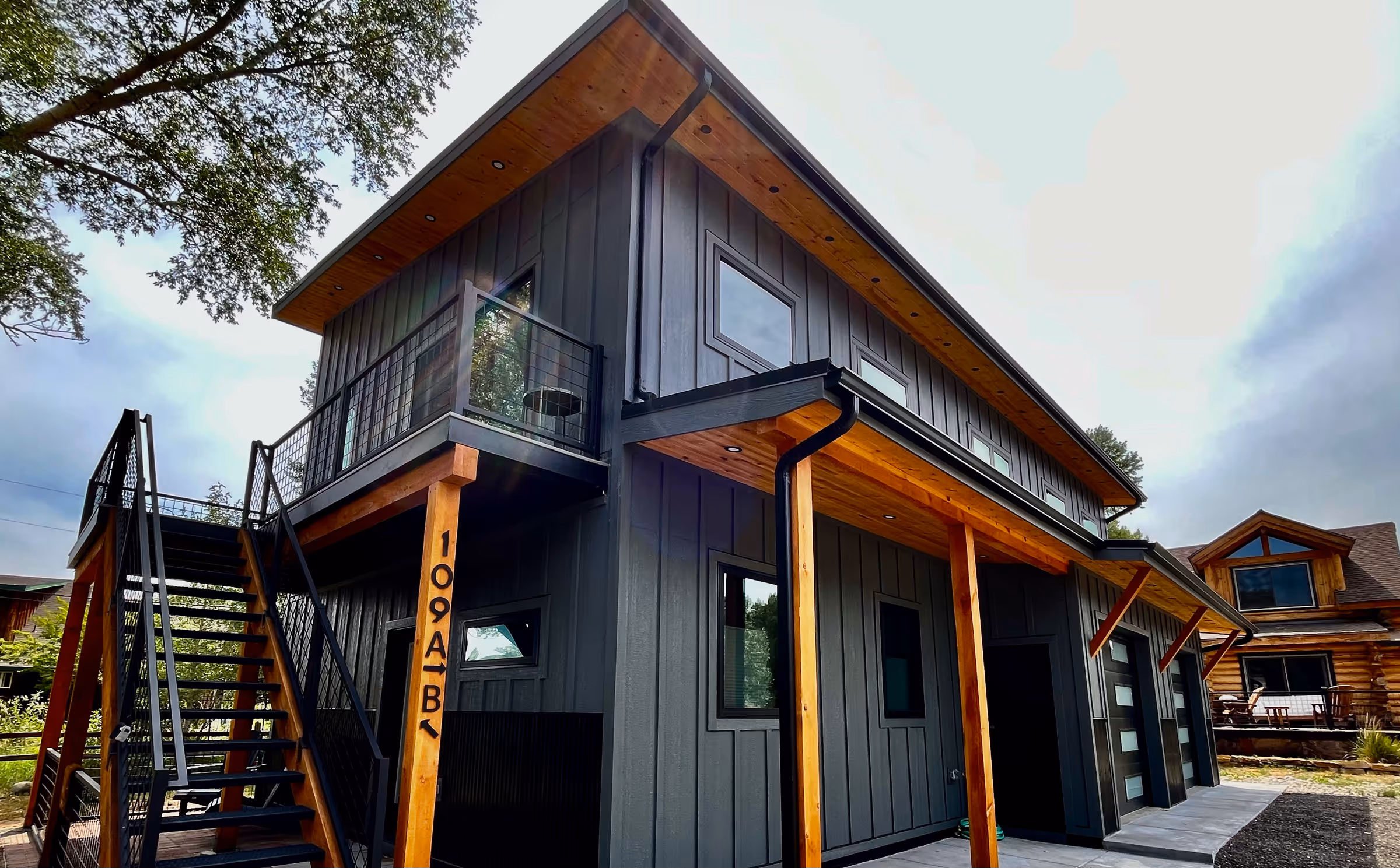 Modern two-story home with dark gray board and batten siding, exposed wood eaves, a black metal staircase leading to an upper balcony, and adjacent rustic log cabin in the background.