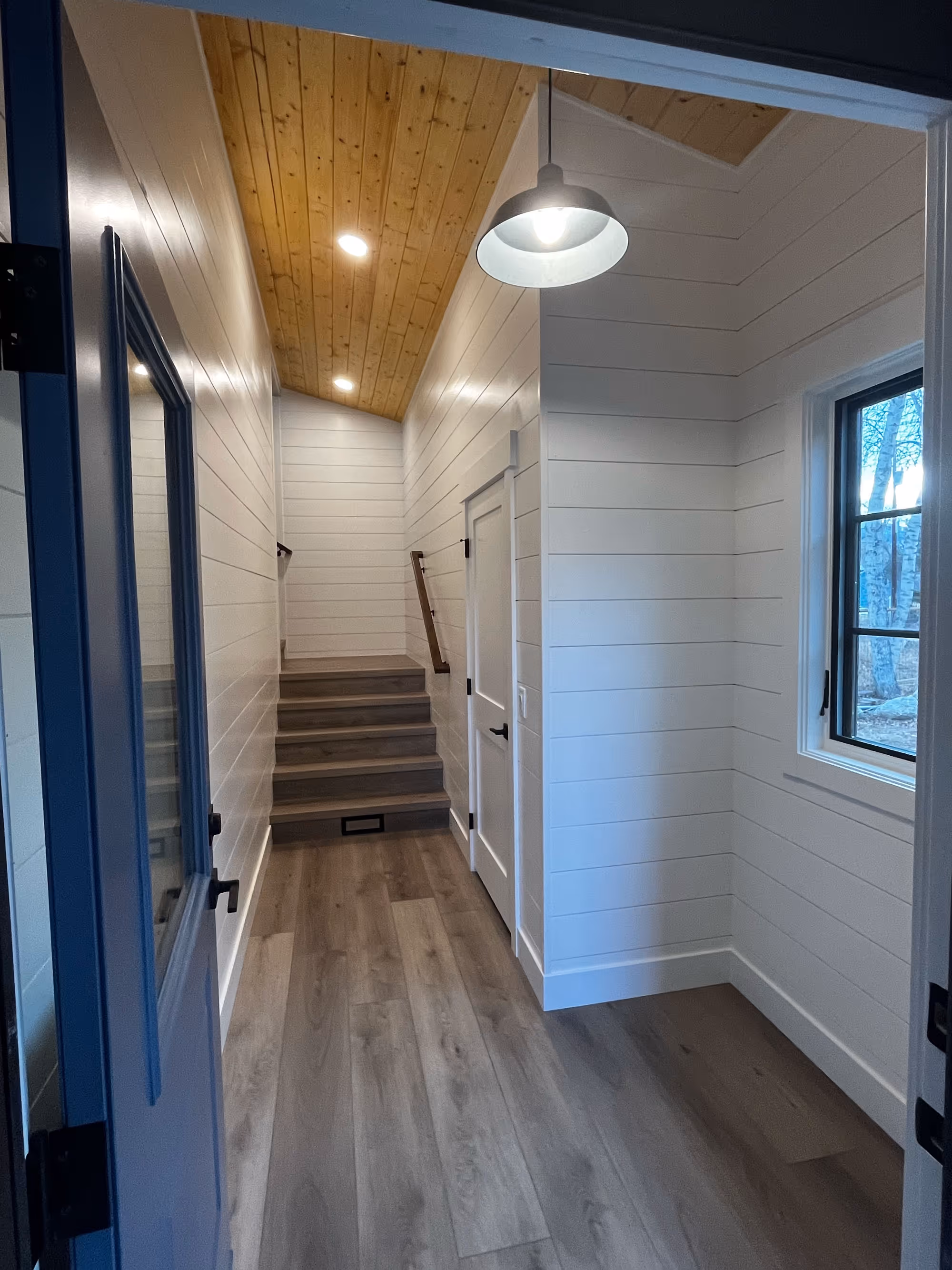 Warm and welcoming home entryway with wood-style flooring, white shiplap walls, wood plank ceiling, and a staircase leading upward from the hallway.