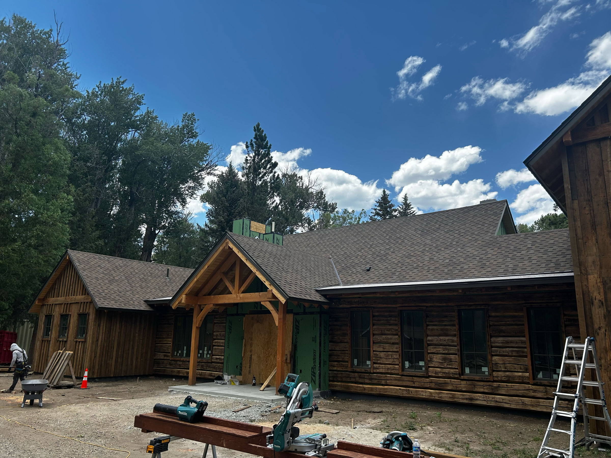 Wood-framed log-style home under renovation with exposed beams, green sheathing, and construction tools visible in the foreground