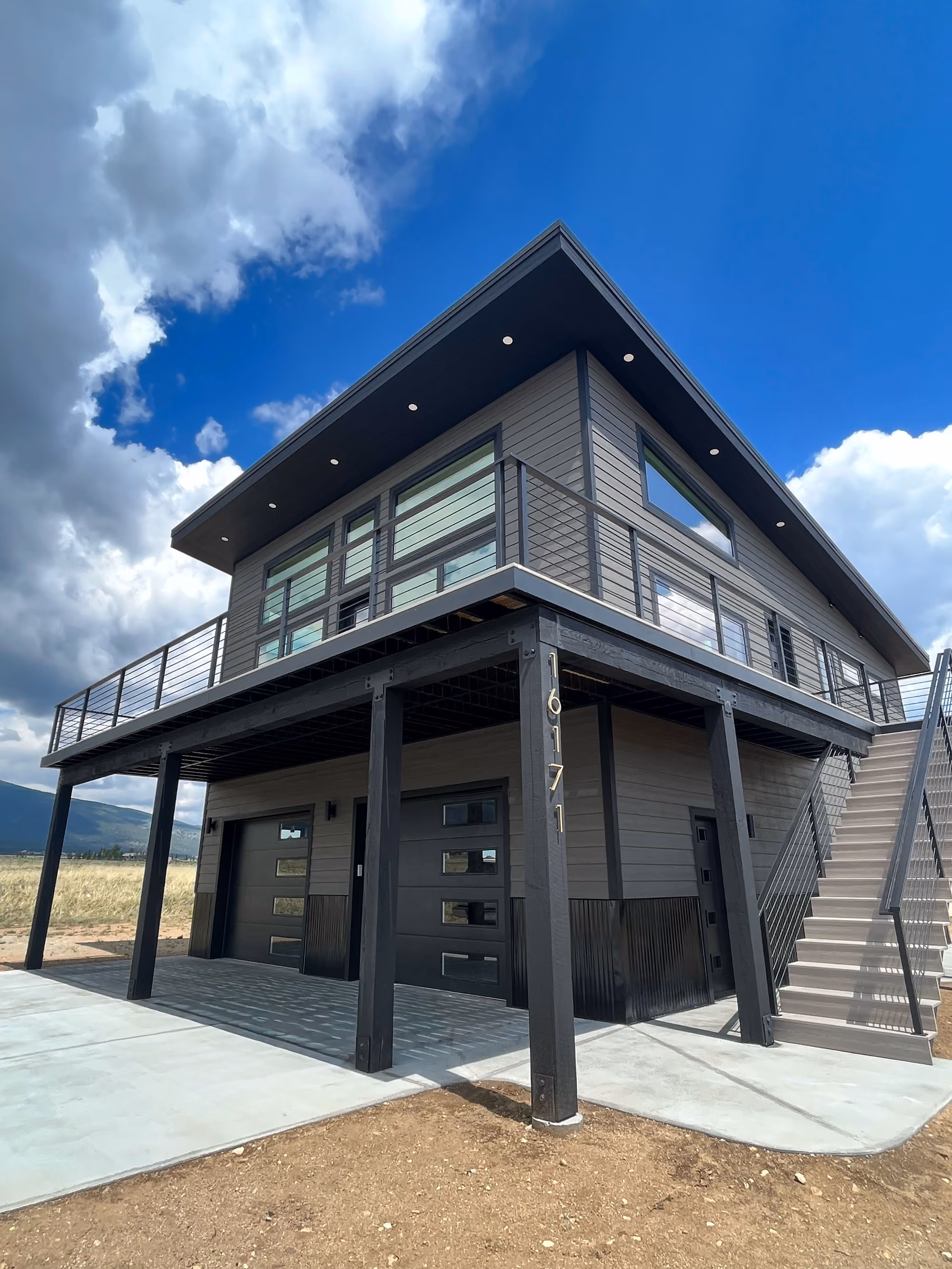 Contemporary two-story home with dark siding, black steel railing, and a sloped roof set against a bright sky with mountains in the distance