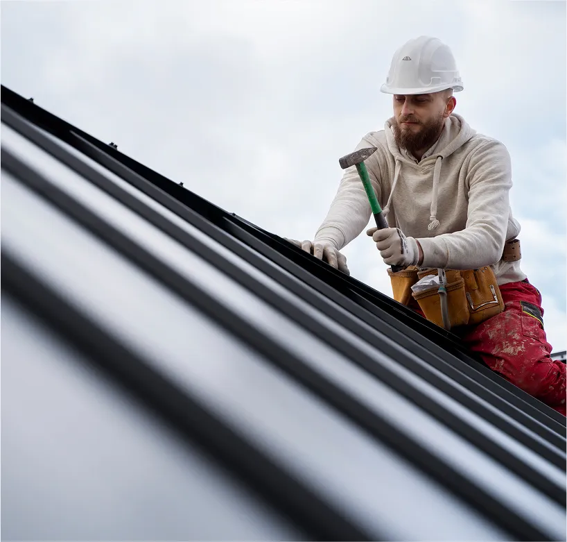 A construction worker wearing a white hard hat and beige hoodie is using a hammer while working on a roof. He is also wearing protective gloves and a tool belt around his waist. The background shows a cloudy sky