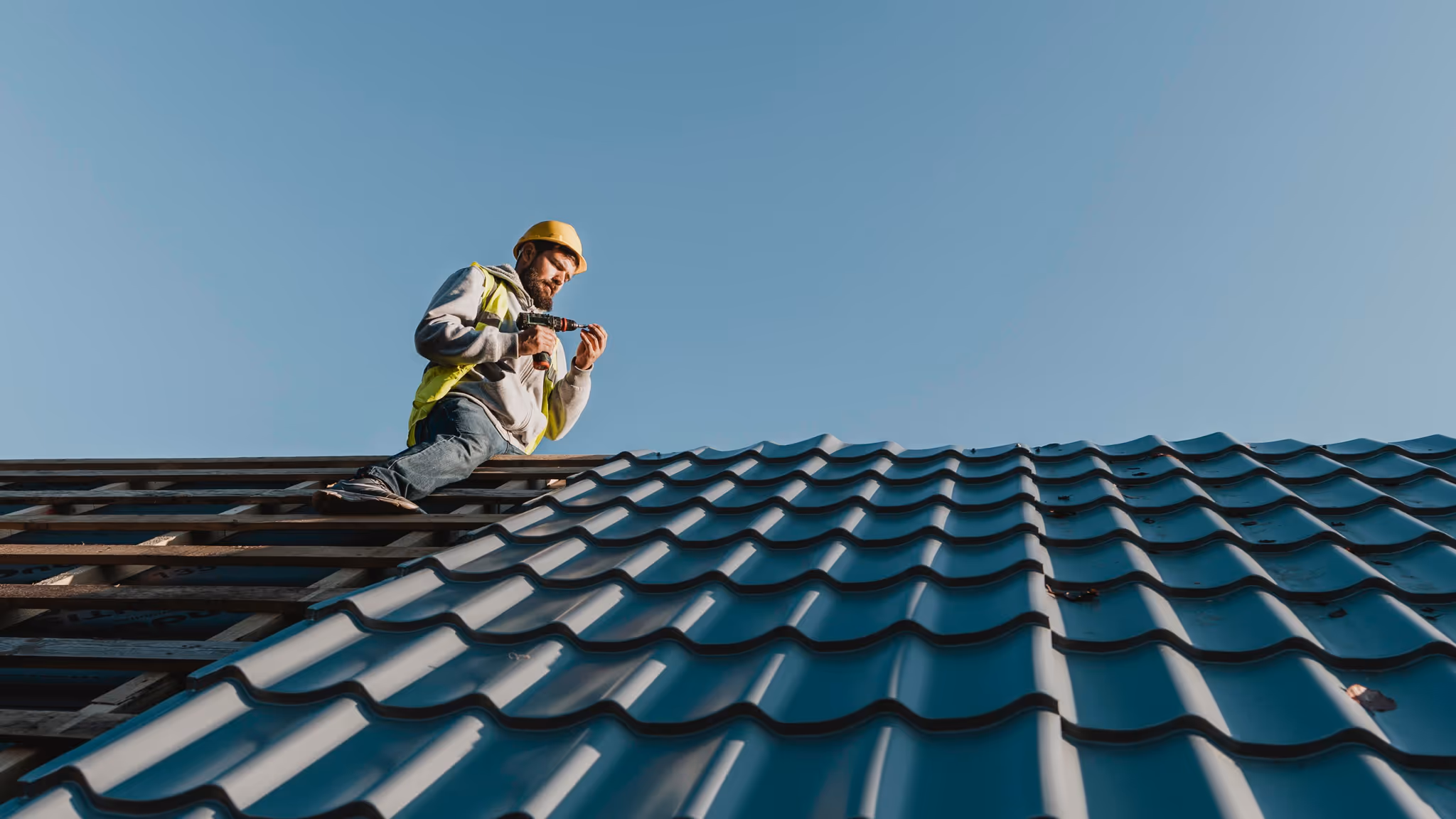 A construction worker wearing a yellow hard hat and reflective vest is sitting on a roof frame while holding a power drill. He appears to be installing or inspecting metal roof tiles under a clear blue sky.