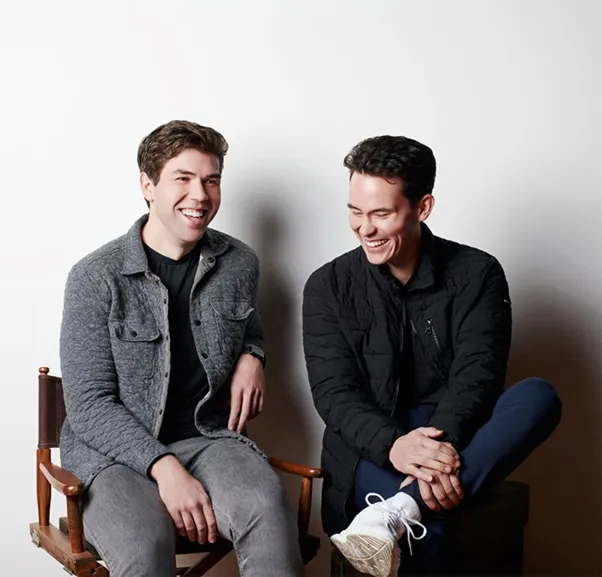 Two men are sitting on chairs and smiling.

