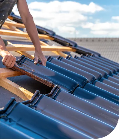 A close-up of a worker’s hands installing dark blue ceramic roof tiles onto a wooden roof frame. The sky is partly cloudy, and rooftops of other buildings are visible in the background.