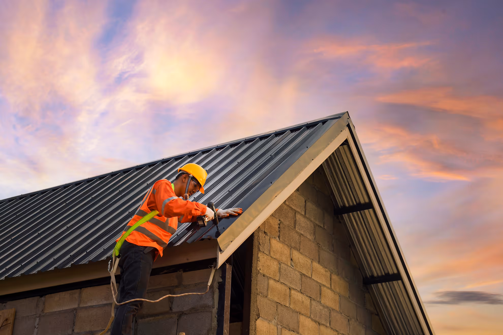 A construction worker wearing a yellow safety helmet and orange reflective vest is standing on a ladder, securing a metal roof panel with a power drill. The building is made of concrete blocks, and the sky in the background is filled with warm, colorful clouds during sunset