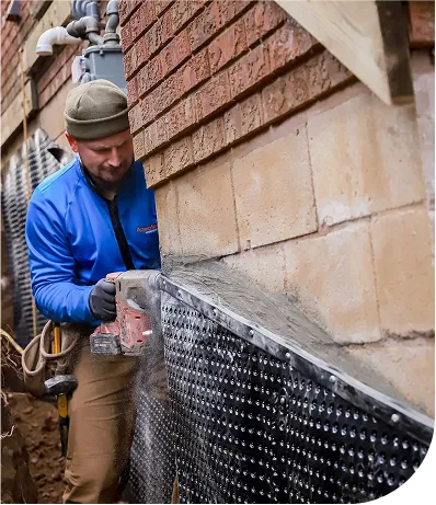 A construction worker in a blue jacket and green beanie is using a power tool to secure a waterproofing membrane to the foundation wall of a brick building. Dust is visible as the tool cuts into the material, and the area appears to be part of a basement or foundation waterproofing project