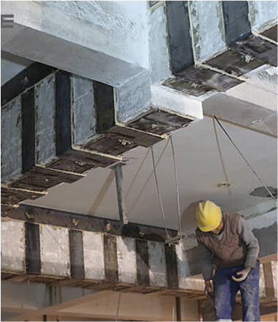 Construction worker reinforcing concrete beams with carbon fiber strips on a building ceiling.