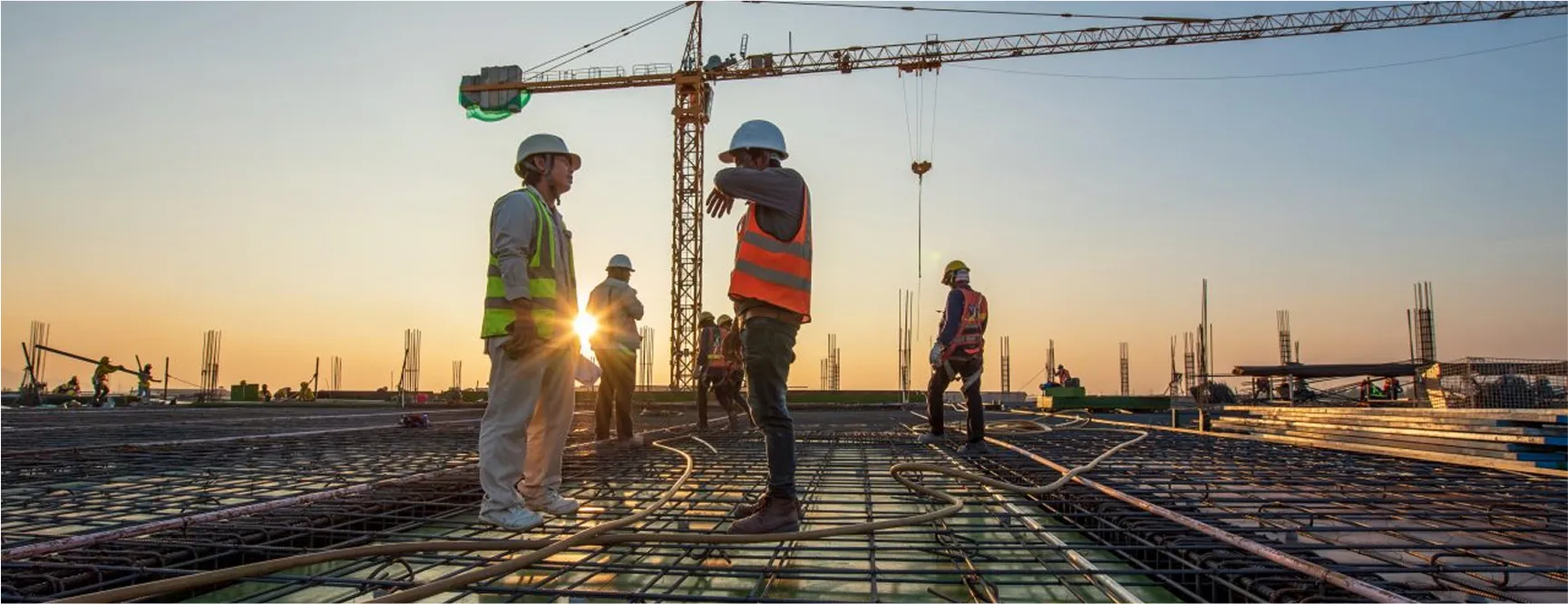 Construction workers preparing steel reinforcement on a high-rise building site at sunset, with a crane in the background.