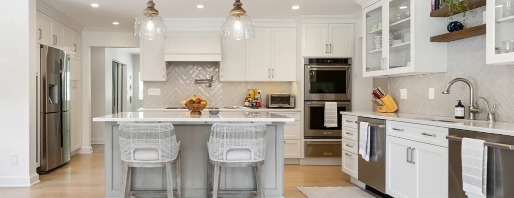 Bright kitchen with white cabinetry, a marble countertop island with gray woven bar stools, glass pendant lights, and stainless steel appliances. The kitchen has light wood flooring and a neutral-colored rug
