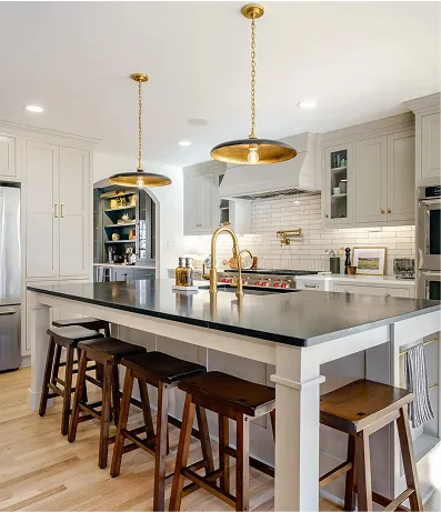 Contemporary kitchen with white cabinets, a black countertop island, gold fixtures including a faucet and pendant lights, and wooden bar stools. The space has light wood flooring and an open layout leading to a living area