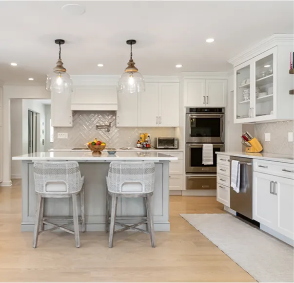 Bright kitchen with white cabinetry, a marble countertop island with gray woven bar stools, glass pendant lights, and stainless steel appliances. The kitchen has light wood flooring and a neutral-colored rug
