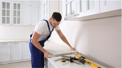 Worker in overalls installing a white countertop in a modern kitchen, using tools for precise fitting along the wall.
