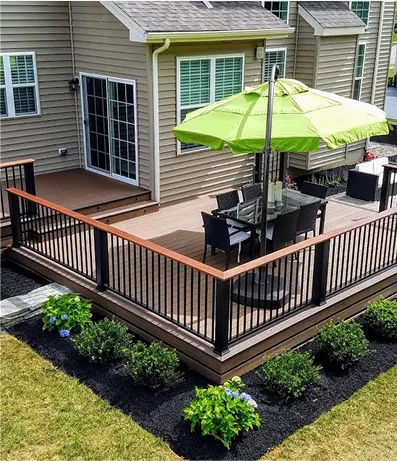 Elevated backyard deck attached to a beige house, featuring a glass dining table with black chairs and a large green umbrella, surrounded by a landscaped garden with neatly trimmed shrubs