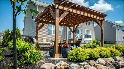 Wooden pergola structure over a stone patio with outdoor furniture, in a landscaped backyard beside a house