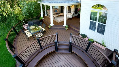 Curved wooden deck attached to a house, featuring white railings and outdoor seating, surrounded by green landscaping.