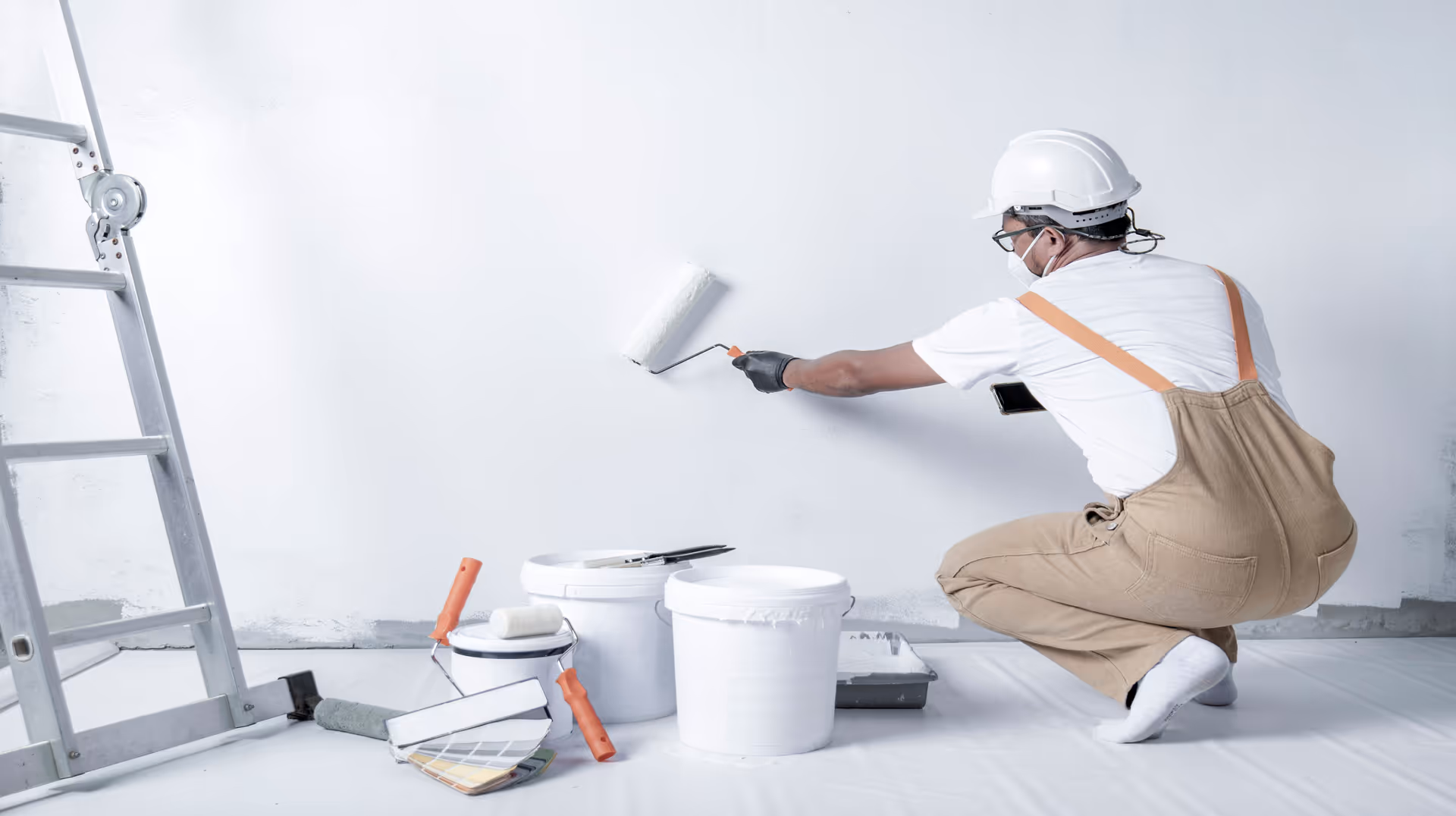 A person wearing a white hard hat, glasses, a white t-shirt, brown overalls, and a face mask is kneeling on the floor while painting a white wall with a paint roller. Several paint buckets, rollers, brushes, a paint tray, a color swatch booklet, and a ladder are nearby. The floor is covered with a protective sheet.