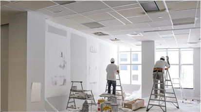 Workers on ladders repairing and preparing white drywall in a bright room.