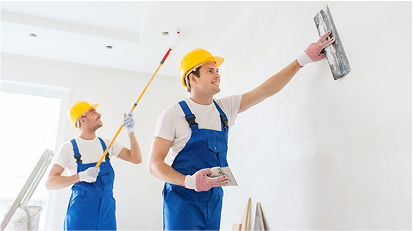 Two workers in blue overalls and yellow helmets painting interior walls with rollers.