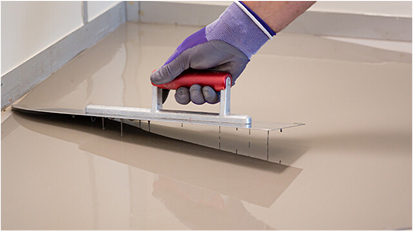Close-up of hand using a trowel to spread leveling compound on a subfloor.
