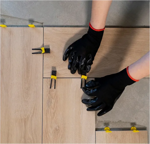 Hands wearing black gloves installing wood-look floor tiles with yellow tile leveling spacers.