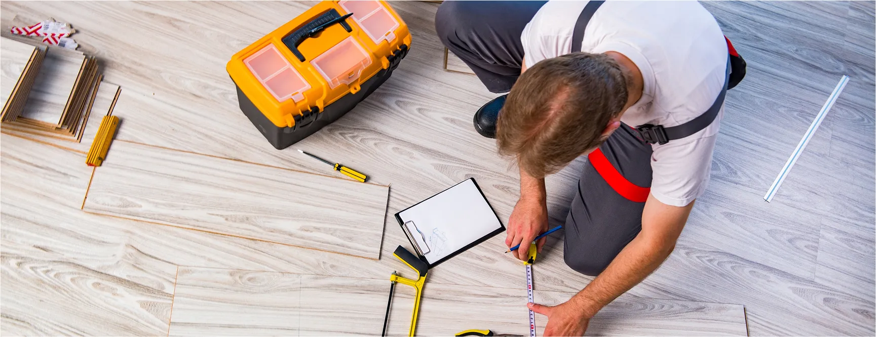 A man kneeling on a wooden floor while measuring a piece of flooring with a tape measure and pencil. Around him are various tools including a yellow toolbox, a screwdriver, a saw, and a clipboard with a paper. He is wearing a white t-shirt with dark pants and red suspenders.