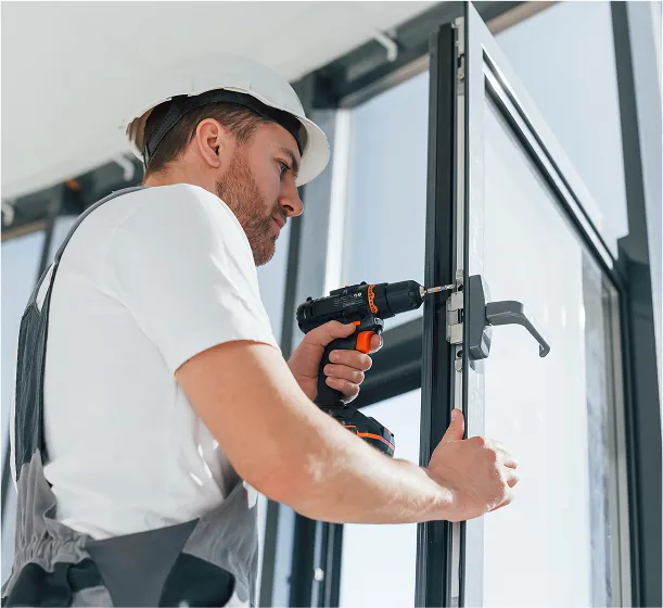 A man wearing a white shirt and a safety helmet is using a power drill to install or repair the frame and handle on glass doors. He is focused on precise work, holding the door with one hand while operating the tool with the other. This image illustrates detailed and professional door or window installation.