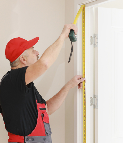 A man wearing a red cap and red overalls is carefully measuring the height of a door frame using a tape measure. He is focused on getting precise measurements, which is an important step in tasks like door installation or fitting. The neutral-colored wall and white door frame provide a clean background, highlighting the attention to detail in the work.
