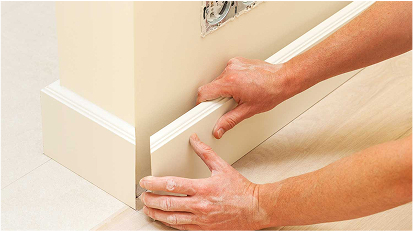 Hands are fitting a white baseboard trim along the bottom of a wall.