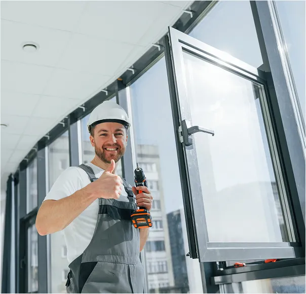 A construction worker wearing a hard hat and overalls is installing a window and giving a thumbs-up with a smile.